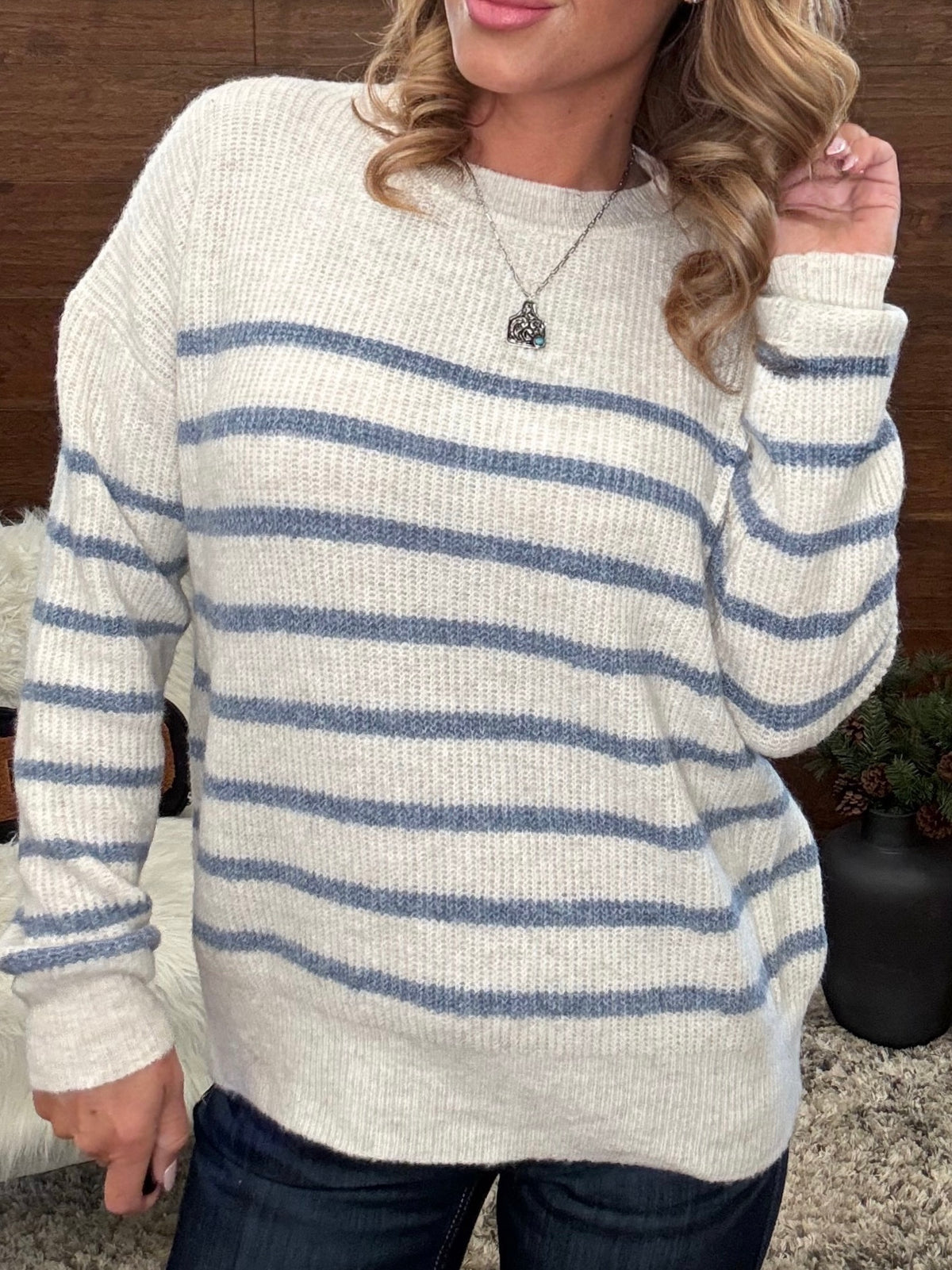 A woman wearing a cream-colored knit sweater with horizontal blue stripes, paired with dark blue jeans and a silver pendant necklace.