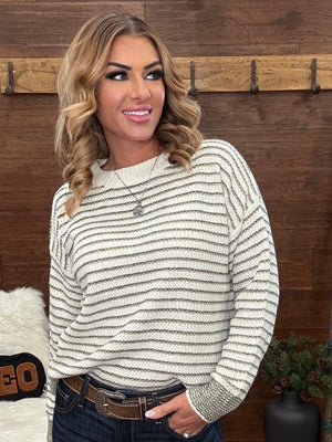 A woman wearing a cream and olive horizontally striped knit sweater, paired with dark blue jeans and a brown belt, standing in front of a rustic wooden wall.