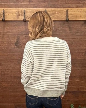 A woman viewed from behind wearing a cream and olive striped knit sweater and dark blue jeans, standing in front of a rustic wooden wall with a coat rack.