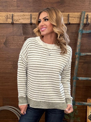 A woman wearing a cream and olive green horizontally striped knit sweater, paired with dark blue jeans, standing in front of a rustic wooden wall with a rustic ladder.