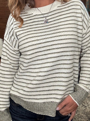 A close-up of a person wearing a cream and green  horizontally striped knit sweater with ribbed cuffs and hem, paired with dark blue jeans.