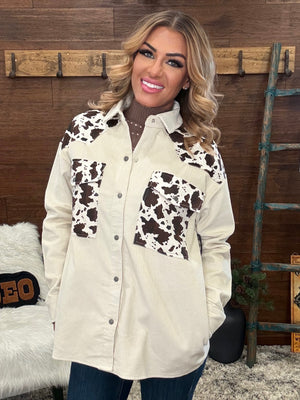 A woman wearing a cream-colored long-sleeve button-up shirt with brown and white cow-print panels on the chest and shoulders, paired with dark blue jeans, standing in front of a rustic wooden wall.