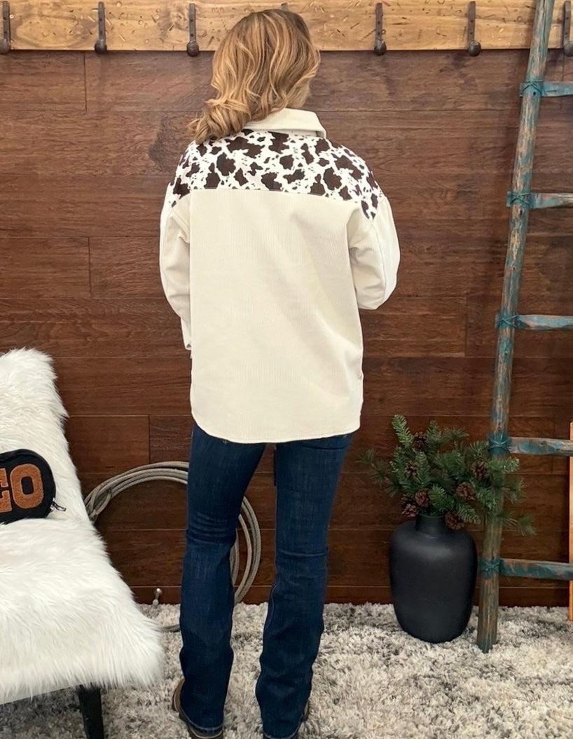A woman stands with her back to the camera wearing a cream western-style shirt with a black and white cow print yoke, paired with dark blue jeans, in front of a rustic wooden wall.