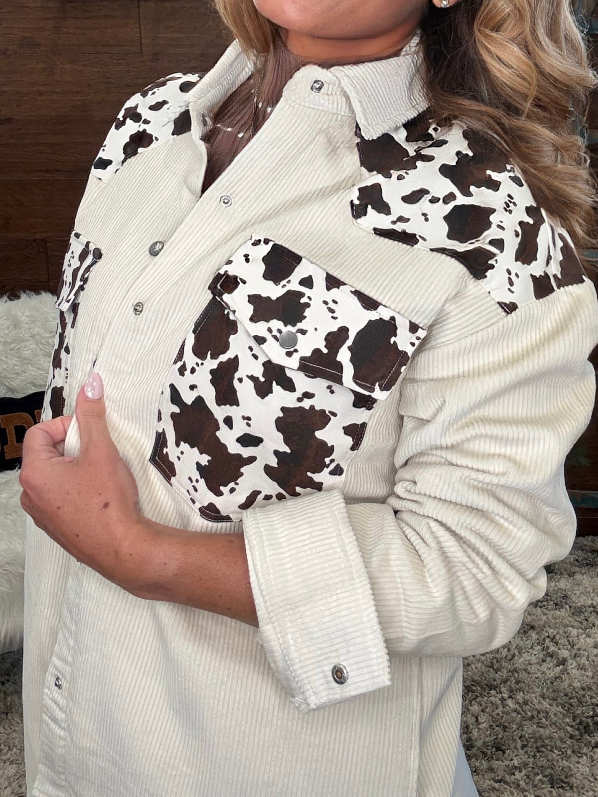 A close-up of a woman wearing a long-sleeve button-front shirt in cream corduroy with brown and white cow-print panels on the chest and shoulders, plus matching cow-print chest pockets.