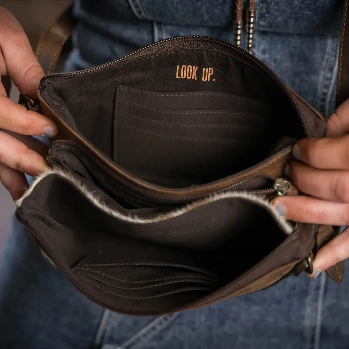 Brown leather bag with 'LOOK UP.' text held by a person wearing blue jeans.