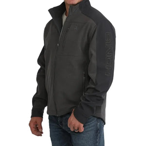 A man wearing a dark gray and black Clinch outdoor jacket with a full-zip front, zippered chest pocket, and vertical Clinch branding on the sleeve, paired with blue jeans against a white background.