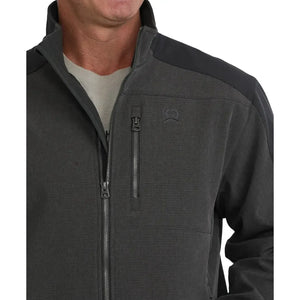 A man wearing a dark gray and black Clinch outdoor jacket with a full-zip front, zippered chest pocket, and vertical Clinch branding on the sleeve, paired with blue jeans against a white background.