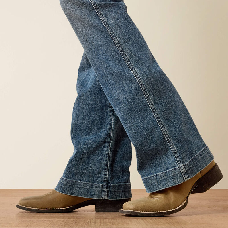 Person wearing blue jeans and brown boots on a wooden floor with a beige wall background