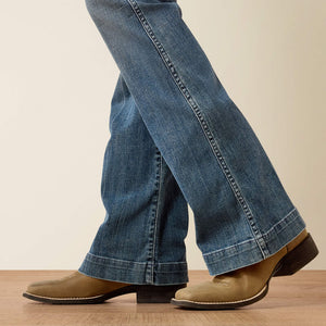 Person wearing blue jeans and brown boots on a wooden floor with a beige wall background