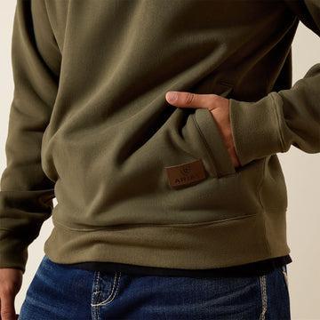 Close-up of a person wearing an olive green sweatshirt with a kangaroo pocket and a brown leather brand patch, paired with blue jeans against a light beige background.