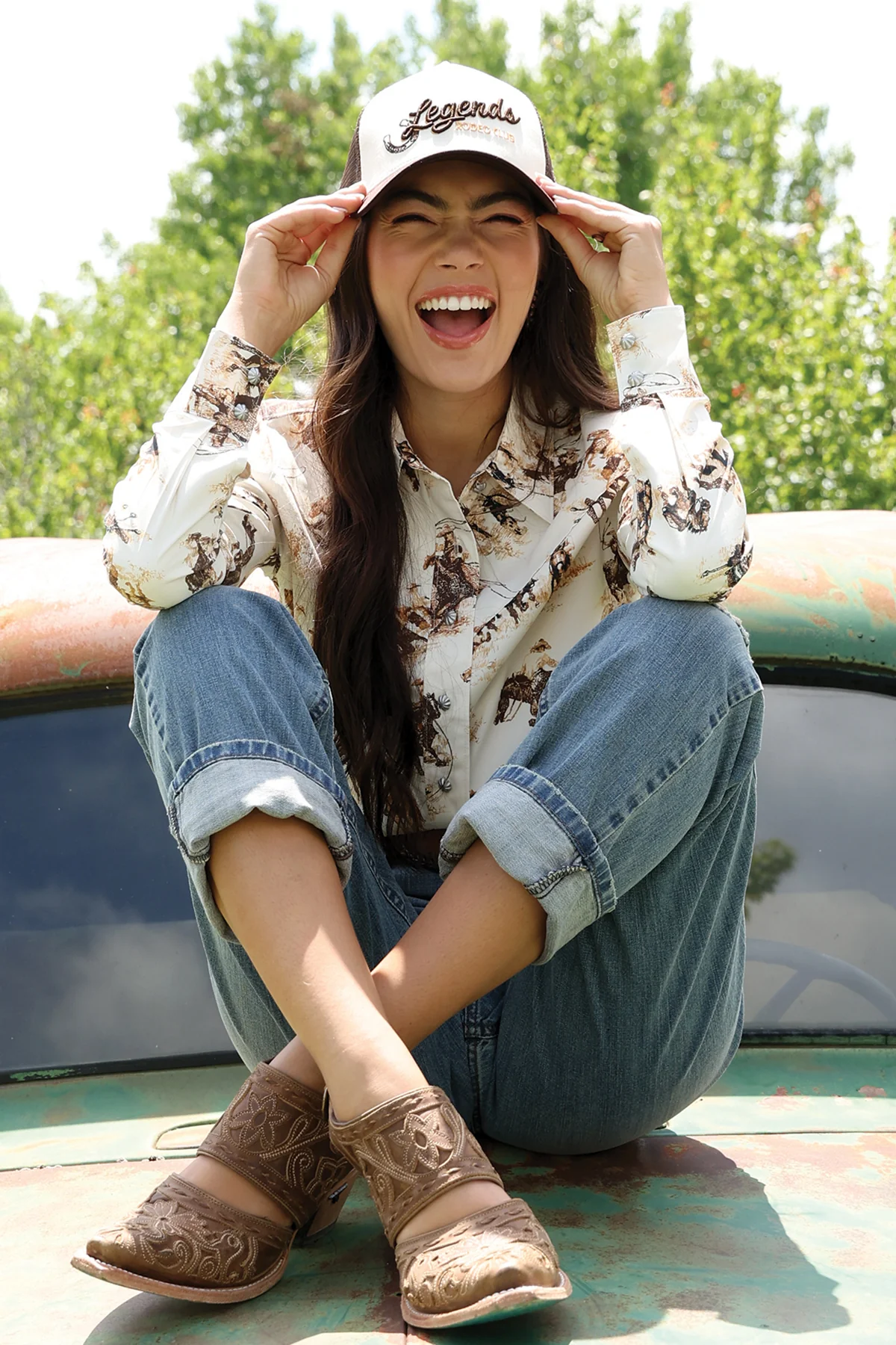 Woman wearing a cap, patterned shirt, jeans, and brown boots sitting outdoors.