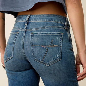 Close-up back view of a person wearing medium-wash blue denim jeans with a distinctive embroidered logo on the back pocket, paired with a blue cropped top against a plain beige background.