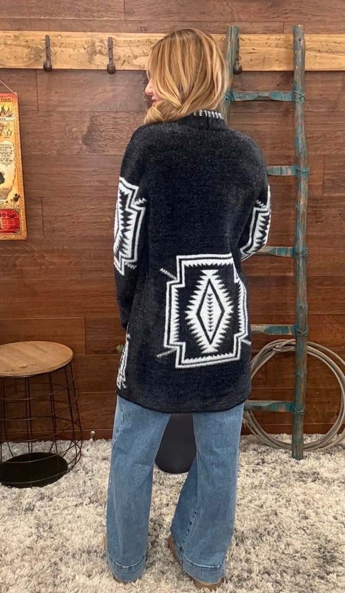 A woman stands with her back to the camera wearing a dark charcoal fuzzy cardigan with large white geometric patterns, paired with wide-leg blue jeans, in a rustic room with a wooden wall and ladder.