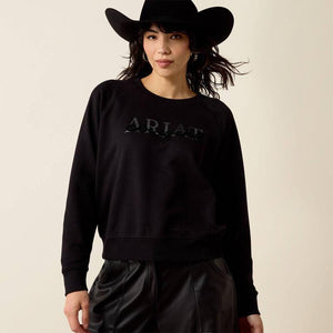 A woman wearing a black crewneck sweatshirt with a tonal black 'ARIAT' logo on the chest. 