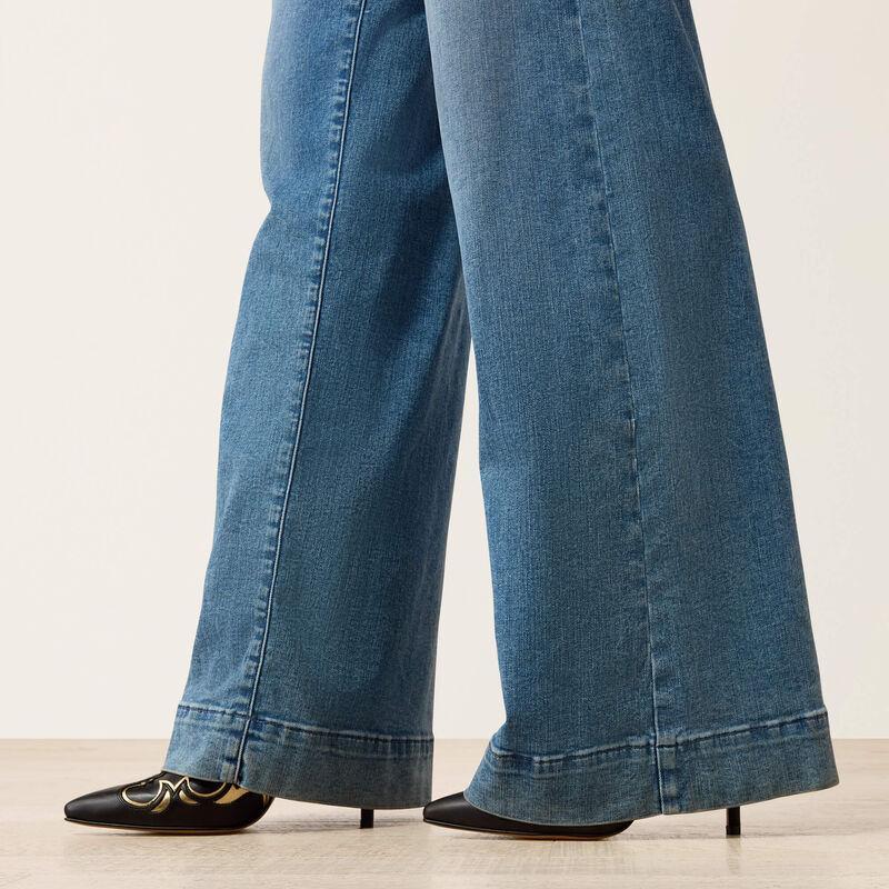 Close-up of a person wearing blue wide-leg jeans with rolled cuffs and black pointed-toe heels against a light neutral studio background.