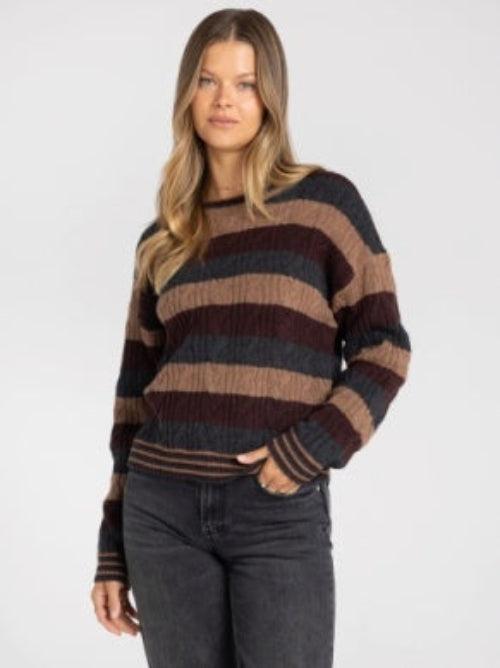 A woman wearing a long-sleeve crewneck sweater with horizontal stripes in dark brown, tan, and charcoal gray