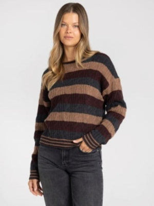 A woman wearing a long-sleeve crewneck sweater with horizontal stripes in dark brown, tan, and charcoal gray