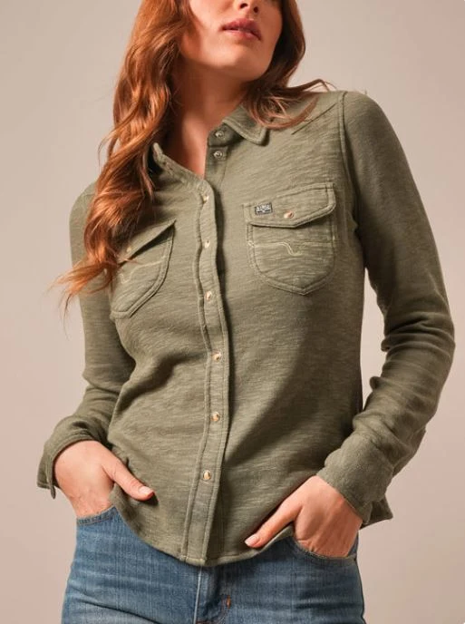 A woman wearing a long-sleeve olive green button-up shirt with two chest pockets, paired with blue jeans, against a plain beige studio background.