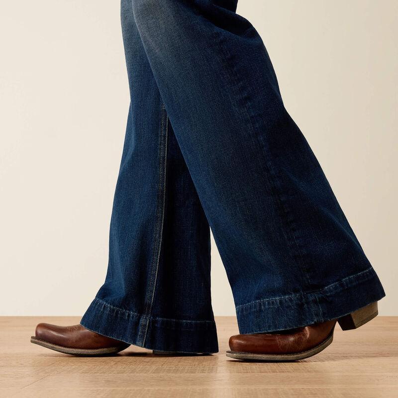 Close-up of a person's legs wearing dark blue flared jeans and brown leather cowboy boots, standing on a light wood floor against a plain beige wall.