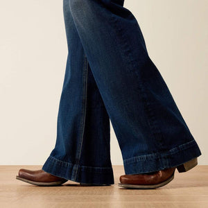 Close-up of a person's legs wearing dark blue flared jeans and brown leather cowboy boots, standing on a light wood floor against a plain beige wall.