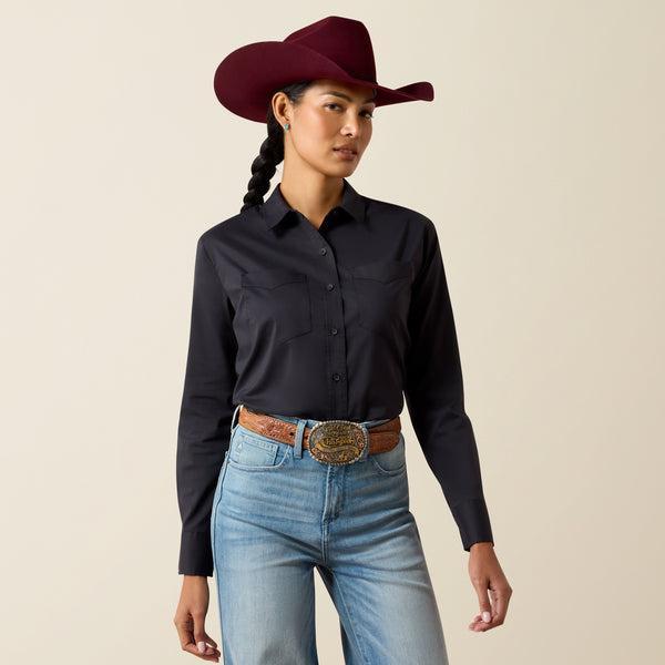 A woman modeling a black long-sleeve western-style button-up shirt, paired with a dark red cowboy hat, blue jeans, and a brown leather belt with a large silver buckle, against a solid beige studio background.