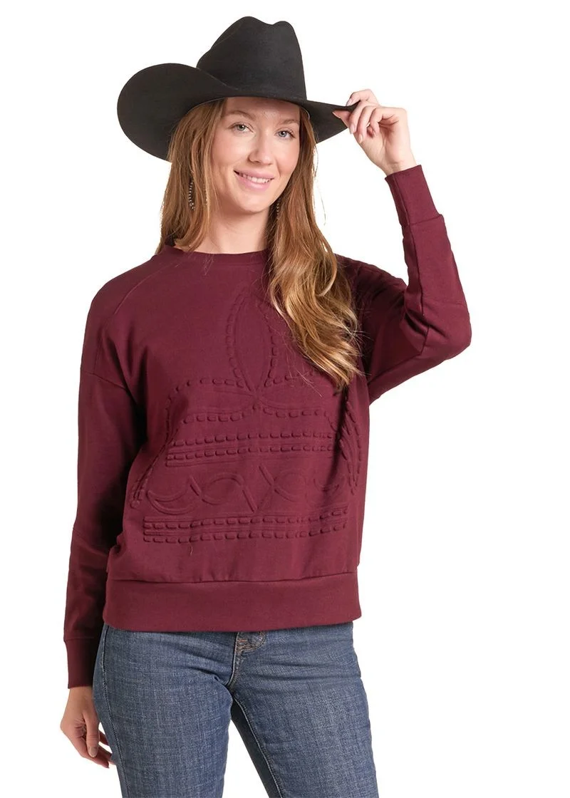 A woman wearing a maroon crewneck sweatshirt with a western-style cable knit pattern, blue jeans, and a black cowboy hat, posing against a white background.