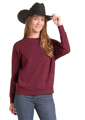A woman wearing a maroon crewneck sweatshirt with a western-style cable knit pattern, blue jeans, and a black cowboy hat, posing against a white background.
