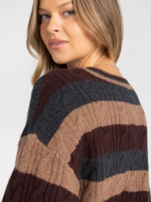 A woman wearing a long-sleeve crewneck sweater with horizontal stripes in dark brown, tan, and charcoal gray