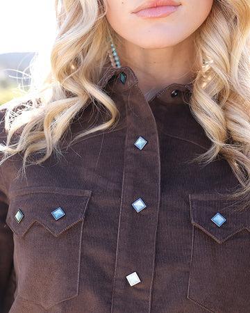 Close-up of a woman wearing a dark brown corduroy button-up shirt jacket with decorative light blue and white diamond-shaped studs on the chest pockets and front placket.
