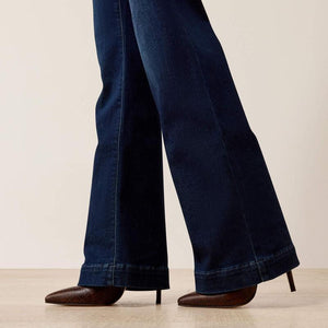 Close-up of a person's legs wearing dark blue wide-leg jeans and brown pointed-toe heels, standing on a light wood floor against a plain beige wall.