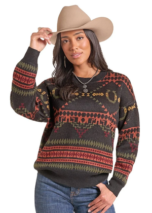 A woman wearing a dark brown knit sweater with a colorful geometric southwestern pattern in red, green, and beige, paired with a tan cowboy hat and blue jeans, set against a plain white background.