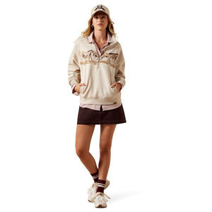 A woman modeling a beige quarter-zip pullover sweatshirt with a mountain graphic across the chest, paired with a dark brown mini skirt, a baseball cap, socks, and sneakers, against a solid white background.