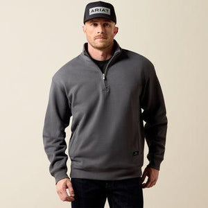 A man wearing a dark gray quarter-zip sweatshirt, a black cap with 'ARIAT' on the front, and dark jeans, standing against a solid beige background.