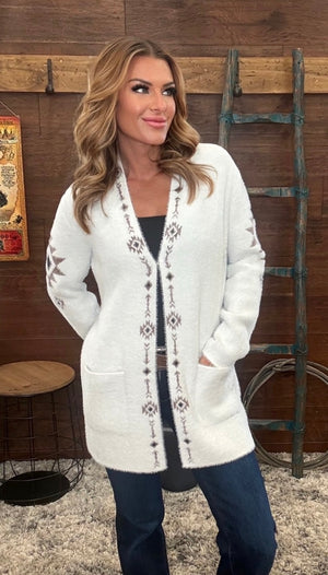 A woman modeling a white open-front cardigan with dark geometric pattern down the front and along the sleeves, paired with a black top and blue jeans, set against a rustic wooden wall backdrop.
