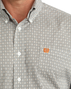 A man wearing a liwhite micro-patterned button-down shirt with a small orange logo on the chest, tucked into blue jeans with a western belt.