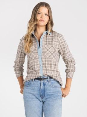 A woman modeling a long-sleeve button-up plaid shirt in beige and brown tones, layered over a light blue collared shirt, paired with light-wash blue jeans against a plain white background.