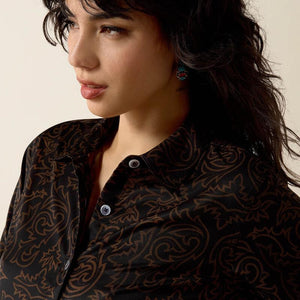 A close-up of a woman wearing a black button-up shirt with a brown paisley pattern, set against a plain beige background.