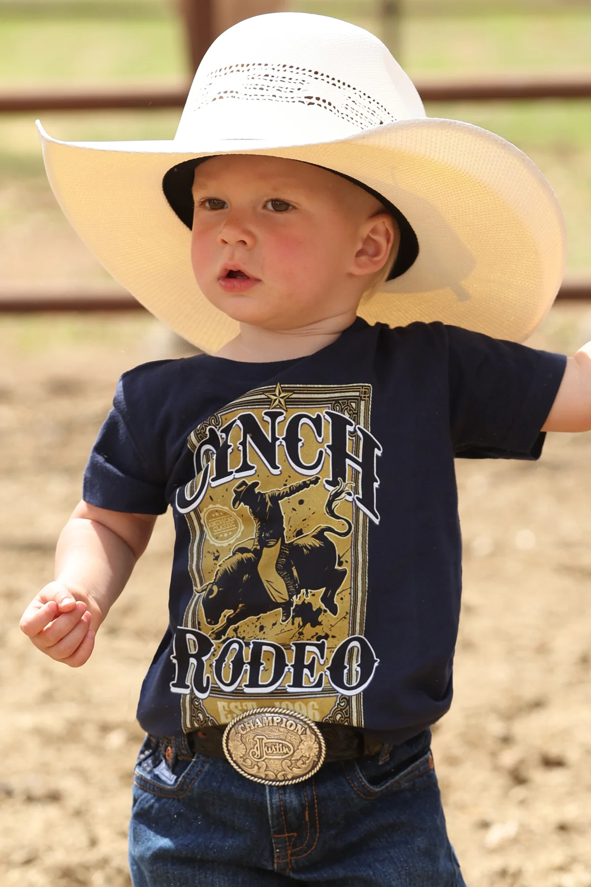 Child wearing a navy blue t-shirt with 'Lunch Rodeo' print, white cowboy hat, and jeans outdoors.