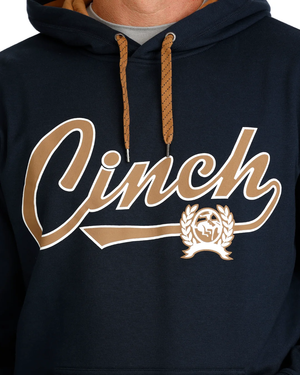 A close-up of a navy blue hoodie featuring a large tan and white 'Cinch' script graphic across the chest, with a matching tan laurel wreath crest underneath. The hoodie has tan drawstrings and is shown on a model.