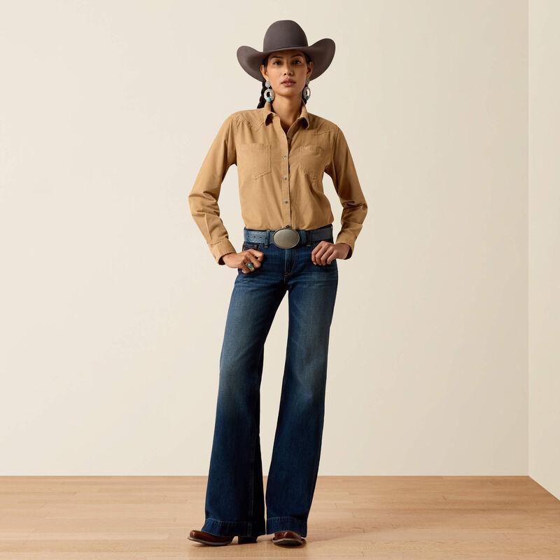 A model stands in a studio wearing a tan western-style button-up shirt, dark blue flared jeans, a brown cowboy hat, and a silver belt with a large round buckle.