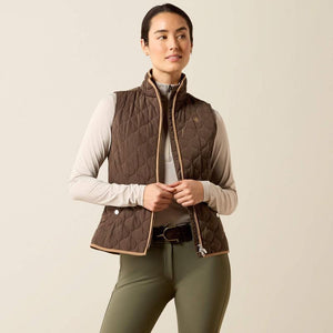 A woman modeling a brown quilted equestrian vest with tan trim, worn over a light beige long-sleeve shirt and olive green riding pants, against a solid light beige background.