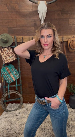 Woman in a black t-shirt and blue jeans posing in a rustic setting with wooden walls and decorative items.