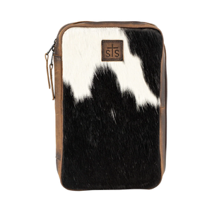 A rectangular zippered pouch featuring a black and white cowhide pattern on the front, with brown leather trim and a brown leather logo patch on the upper right side. The patch displays the 'sts' brand name with a cross.