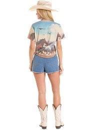 Woman wearing a graphic t-shirt, denim shorts, and cowboy boots with a scenic background.