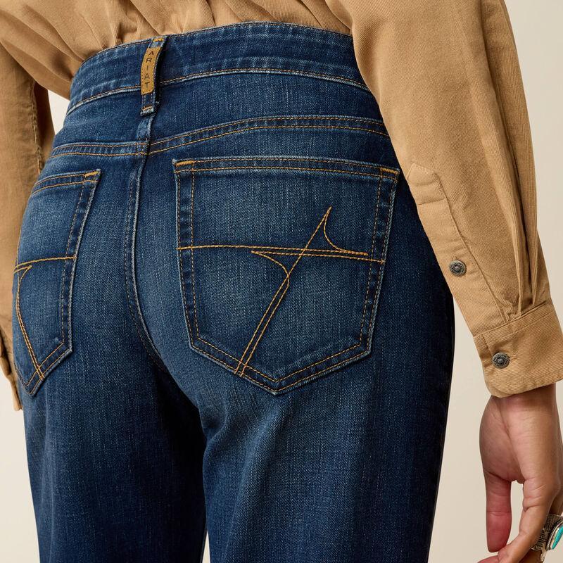 Close-up back view of a person wearing dark blue denim jeans with contrast gold stitching and a tan long-sleeve shirt, against a light beige background.