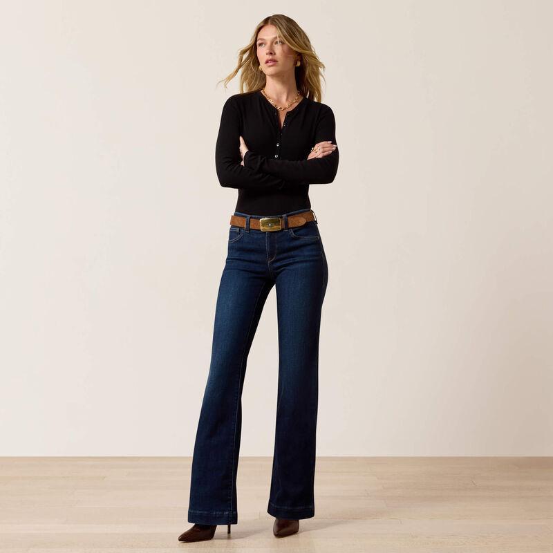 A woman stands in a studio wearing a black long-sleeve henley top, dark blue flare jeans with a brown belt, and brown pointed shoes against a plain light beige wall and pale wood floor.