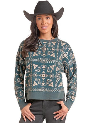 A woman wearing a long-sleeve crewneck sweatshirt with a teal and beige southwestern geometric pattern, paired with a black cowboy hat and dark gray jeans, against a plain white background.