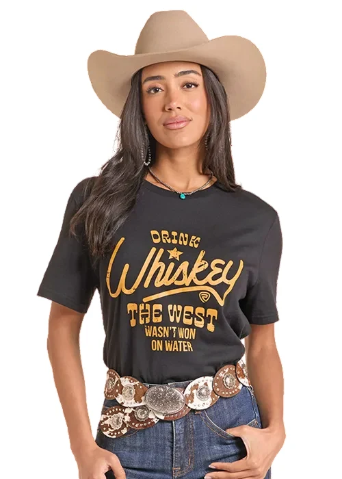 A woman wearing a tan cowboy hat, a black graphic t-shirt with gold lettering that reads 'DRINK Whiskey THE WEST WASN'T NOW ON WATER', blue jeans, and a large silver concho belt, posed against a solid white background.