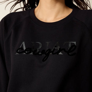 A woman wearing a black crewneck sweatshirt with a tonal black 'ARIAT' logo on the chest. 