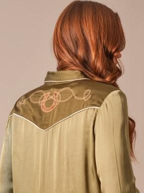 A woman viewed from behind wearing a satin-like sage green shirt jacket with white piping and embroidered western-style lettering across the shoulders.